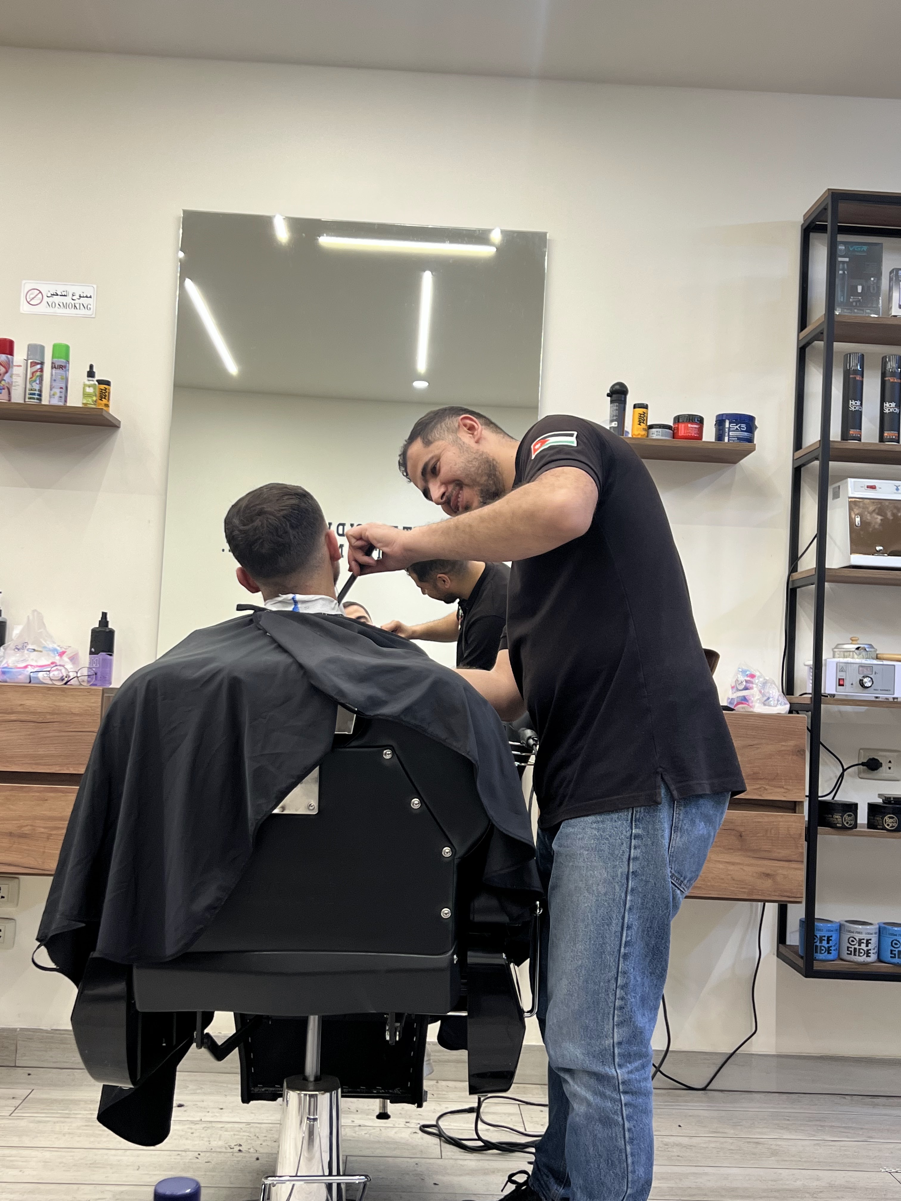 Barber at work on a client, Jordan flag patch on his sleeve, product wall in the background